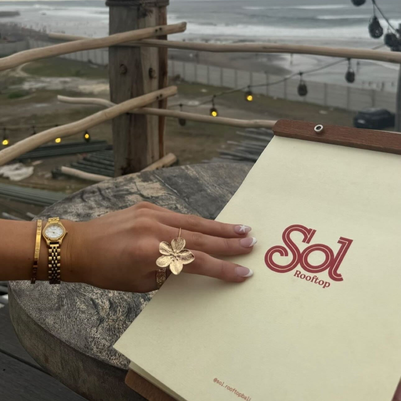 Person's hand wearing the Fleur Ring, a stunning flower statement ring by ÈRE, while holding a menu at a rooftop venue, showcasing its elegance in lifestyle settings.